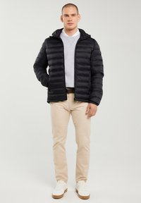 Hombre de pie de frente vistiendo una chaqueta acolchada negra, sudadera gris claro, pantalones beige y zapatillas blancas sobre un fondo claro y liso.