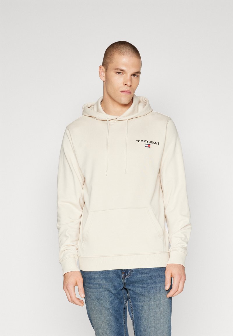 Felpa beige con cappuccio e tasca anteriore, con logo "TOMMY JEANS" ricamato. Abbinata a jeans in denim blu. Design semplice.