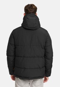 Mand iført sort pufferjakke med hætte, set fra ryggen, stående mod en helt hvid baggrund.