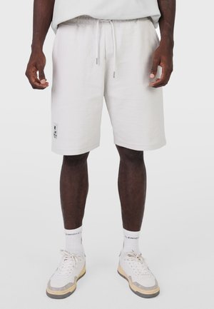 Mężczyzna noszący białe teksturowane szorty z małą naszywką na lewej nodze, w połączeniu z białymi skarpetkami i białymi trampkami.