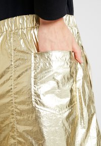 Gonna in tessuto dorato stropicciato con vita elasticizzata. Presenta una tasca laterale e una texture lucida, che mette in evidenza il suo aspetto metallico.