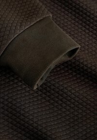 Donkerbruin gewatteerd stof met een close-up van een bijpassende geribbelde gebreide boord die over het getextureerde oppervlak is gevouwen.