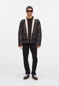 Chaqueta de cuero marrón con cuello de borrego, cierre de cremallera y bolsillos frontales; combinada con un suéter de punto marrón y pantalones negros. Zapatillas negras.