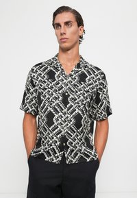Man with slicked-back hair wearing a black and white tropical pattern short-sleeve button-up shirt and dark pants with hands in pockets.