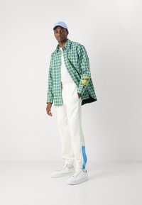 Grønne og blå ternede skjorte, hvid T-shirt, hvide bukser med blå og grønne sidelister, og hvide sneakers. Lys blå kasket.