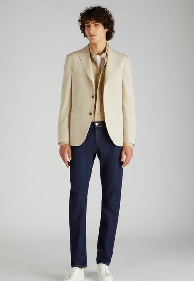Blazer a righe beige con revers a punta, due tasche frontali, bottoni marroni; abbinato a una camicia beige, jeans blu scuro e sneakers bianche.