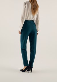 Pantalon sarcelle à coupe ajustée, avec une taille mi-haute, fermeture éclair à la cheville, associé à un chemisier blanc et des talons noirs.