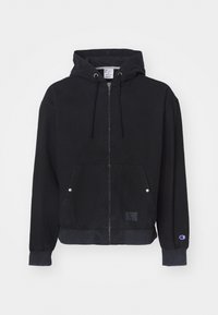 Črna zip-up kapuca iz mehkega materiala, ki ima kenguru žep, rebraste manšete in majhno oznako z logotipom spredaj.