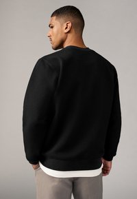 Schwarzer Sweatshirt mit Struktur, mit einem gerippten Rundhalsausschnitt und Bündchen, über einem weißen Shirt getragen. Das Design hat einen lockeren Schnitt und minimale Details.