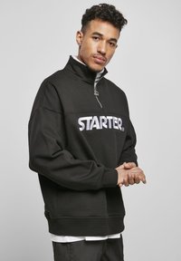 Svart fleece genser med høy krage og halv glidelås. Har en fremtredende hvit "STARTER" logo over brystet. Casual passform.