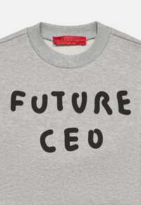 Pilkas megztinis su apvalia iškirpte, ant priekinės pusės išdidžiai parašyta juoda spalva „FUTURE CEO“. Etiketėje parašyta „MAX&Co.“