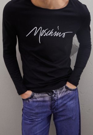 Osoba ubrana w czarną koszulkę z długim rękawem z białym napisem "Moschino" i niebieskie dżinsy, ręce schowane w kieszeniach, na tle szarej ściany.