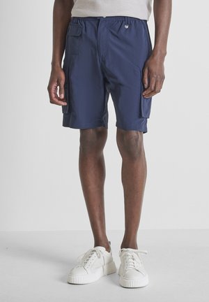 Homme portant un short cargo bleu marine et des baskets blanches, debout devant un fond clair uni.