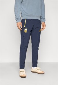 Σκούρες μπλε φόρμες με στενή εφαρμογή, που διαθέτουν χρυσαφένιες λεπτομέρειες, λογότυπο AFA και πλαϊνές τσέπες. Κατασκευασμένες από ελαφρύ υλικό με λεία υφή.