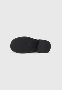 Semelle de sandale noire avec surface en caoutchouc texturé, profil plat, forme contournée et section de talon lisse. Design minimal, sans marquage visible.