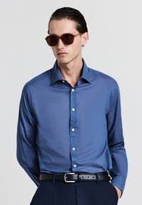 Blaues Langarmhemd mit glatter Textur, durchgehenden Knöpfen und spitzem Kragen. Kombiniert mit runden braunen Sonnenbrillen und einem schwarzen Gürtel.