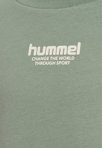 Zielona koszulka bawełniana z białym napisem "hummel" oraz "ZMIENIAJ ŚWIAT PRZEZ SPORT", z okrągłym dekoltem i krótkimi rękawami.