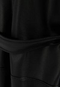 Svart lädertextil med en slät yta och subtil lyster. Har ett midjebälte som är sytt i designen, vilket förbättrar strukturen.