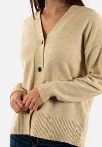 Cardigan lavorato a maglia beige con scollatura a V, due bottoni neri, polsini e orlo a costine, tessuto strutturato e vestibilità ampia.