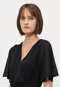 Black dress with a V-neck and short flutter sleeves. Made of lightweight fabric, features a fitted waist and soft draping.