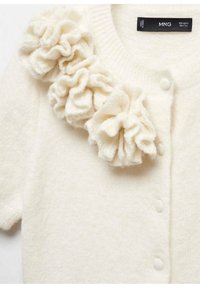 Cardigan en maille crème avec un accent texturé et volanté sur l'épaule gauche, fermé par cinq petits boutons sur le devant.