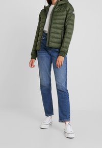 Grüne Puffjacke mit Kapuze, ausgestattet mit einem Reißverschluss und gerippten Bündchen, kombiniert mit blauen Jeans und weißen Sneakern.