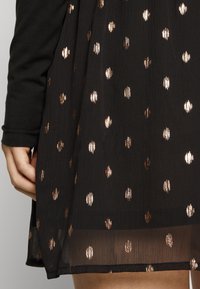 Svart klänning med glänsande kopparprickar. Tyget verkar lätt och har en strukturerad yta. Kjolens kant har en lätt genomskinlig detalj.