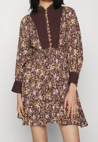 Robe fleurie marron avec des fleurs jaunes et blanches. Dotée d'un corsage texturé, d'une fermeture à boutons et d'un ourlet à volants. Manches longues avec poignets.