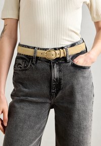 Ceinture en daim beige avec une boucle ronde en or, portée avec un jean noir vieilli taille haute, présentant de grandes poches et une texture légèrement usée.