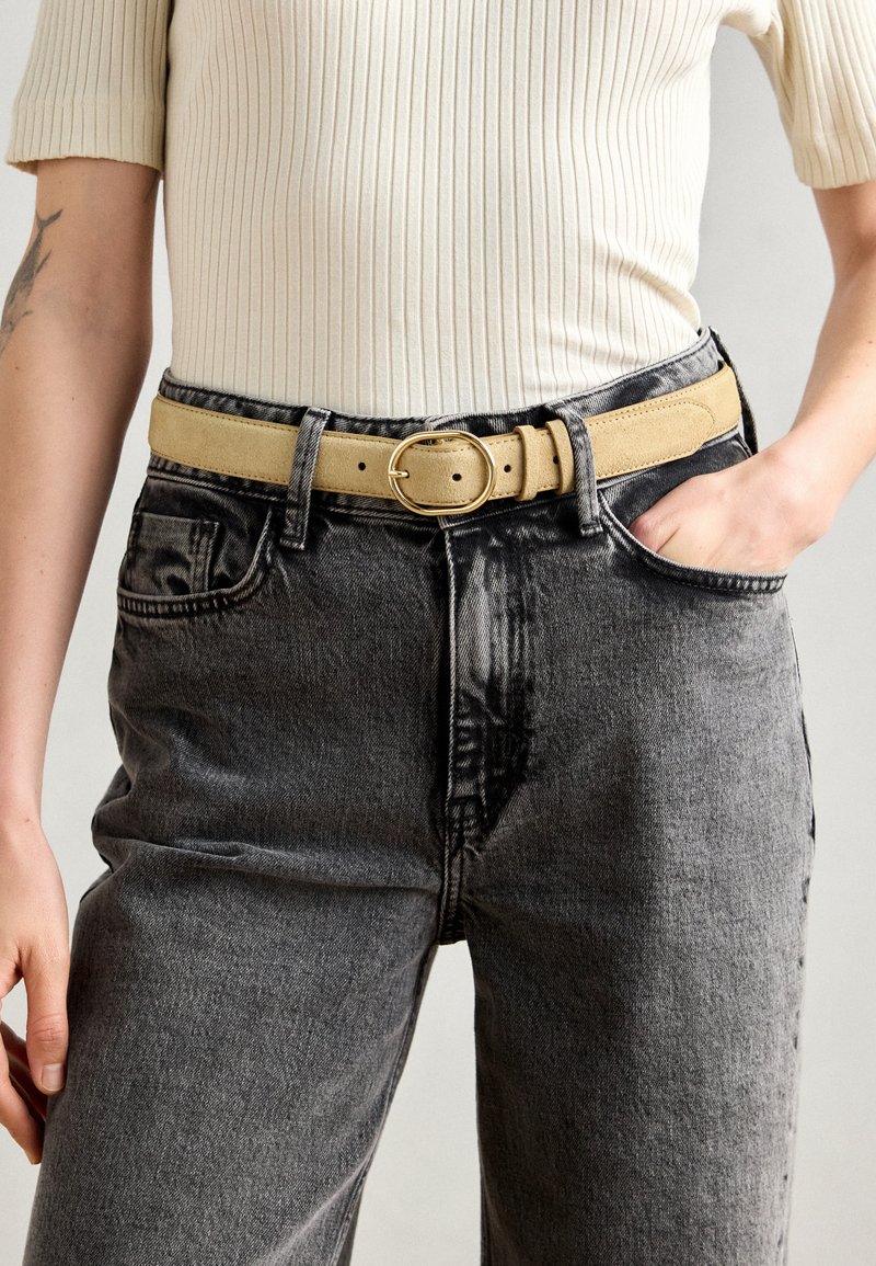Ceinture en daim beige avec une boucle ronde en or, portée avec un jean noir vieilli taille haute, présentant de grandes poches et une texture légèrement usée.