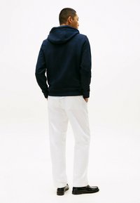 Granatowy hoodie z kieszenią na sznurku, luźny krój i ściągacze, zestawiony z białymi spodniami i czarnymi butami, sfotografowany od tyłu.