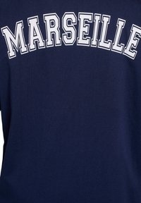 Tamsiai mėlyna marškinėliai su žodžiu "MARSEILLE" paryškintomis baltomis didžiosiomis raidėmis per krūtinę. Lygaus paviršiaus, trumpomis rankovėmis, paprasto dizaino.