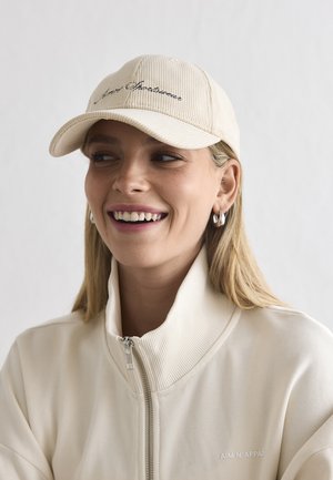 Glimlachende vrouw die een crèmekleurig jack met ritssluiting draagt en een bijpassende corduroy baseballpet met kleine oorringen tegen een effen achtergrond.