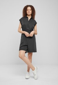 Combinaison noire à manches courtes avec un devant boutonné, un col et des shorts à revers. Portée avec des baskets blanches, présentant une texture de tissu lisse.