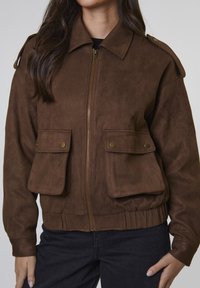 Brązowa skórzana ramoneska z zamkiem błyskawicznym, dwoma przednimi kieszeniami z klapką oraz elastycznym pasem. Posiada pagony na ramionach dla większego efektu.