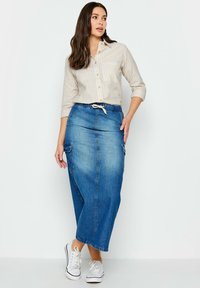 Maxi-Jeansrock in Mittelblau mit einem Kordelzugbund, seitlichen Taschen und einer fließenden Silhouette. Kombiniert mit einem gestreiften Shirt in Weiß und Beige.