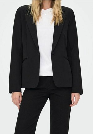 Femme portant un blazer noir et un pantalon noir sur une chemise blanche, debout devant un fond uni, mains détendues le long du corps.