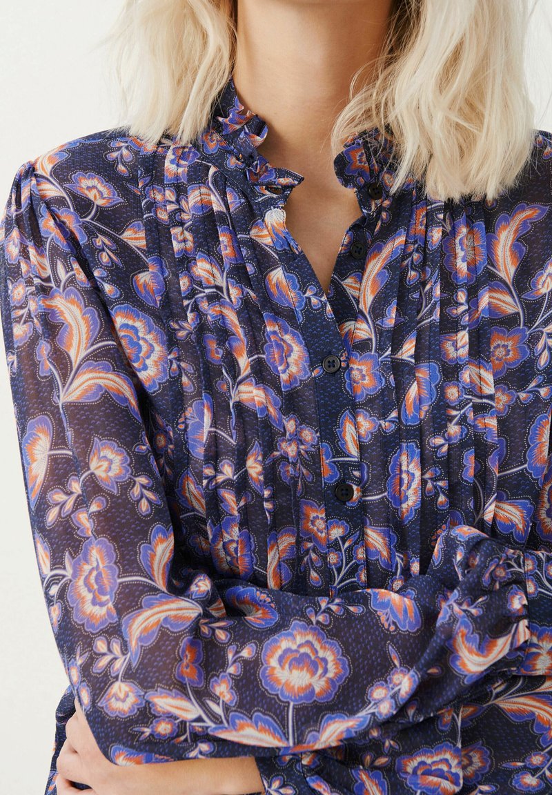 Przezroczysta, ciemnoniebieska bluzka z wyrazistymi wzorami kwiatowymi w kolorze pomarańczowym i różowym. Posiada plisowania, falbaniasty kołnierzyk i czarne guziki.