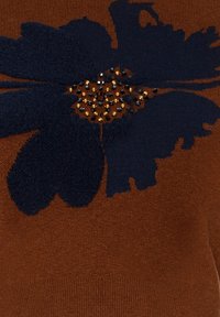 Motif floral bleu marine foncé avec des pétales texturés et de petites perles noires et dorées ornant un tissu tricoté couleur rouille.