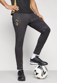 Μαύρο αθλητικό παντελόνι με υφή επιφάνειας, χρυσό λογότυπο και λεπτομέρειες με πέντε αστέρια, συνδυασμένο με μαύρα ποδοσφαιρικά παπούτσια σε μπάλα με σχέδιο.