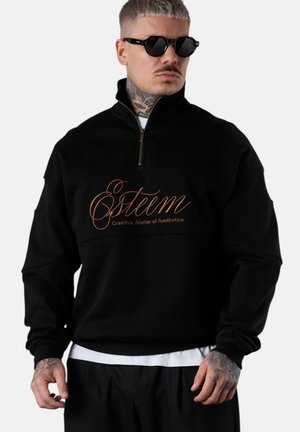 Mann mit Tattoos und Sonnenbrille trägt ein schwarzes Sweatshirt mit Reißverschluss und braunem Text "Estéem Creative Atelier of Aesthetics" sowie schwarze Hosen.
