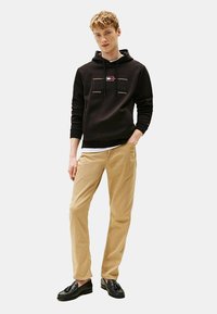 Sort hættetrøje med logo foran, parret med beige bukser. Hættetrøjen har snoretræk og ribbede manchetter, mens skoene er sorte loafer.