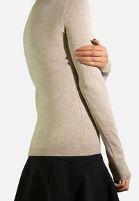 Pull en tricot beige à manches longues avec des poignets et un ourlet côtelés, présentant une coupe slim et une texture douce. Porté sur une jupe sombre.