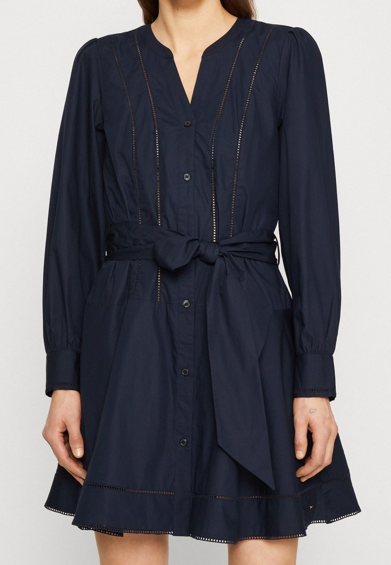 Abito navy con bottoni, maniche lunghe, vita cintrata e cintura annodata. Presenta dettagli a taglio lungo il davanti e l'orlo. Tessuto morbido.
