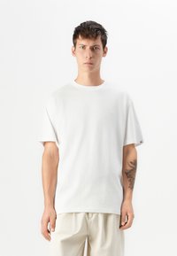 T-shirt branca de algodão com mangas curtas, gola redonda e um logótipo discreto no peito. Combinada com calças de tom claro. Design minimalista.