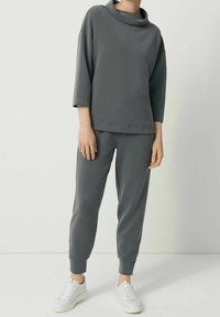 Person som bär en mörkgrå långärmad tröja och matchande avsmalnande byxor med uppvikta anklar, tillsammans med vita sneakers, står på en vit golv.