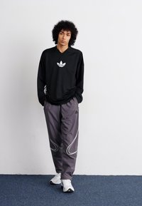 Mustapaitainen pitkähiha, jossa on valkoinen Adidas-logo, yhdistettynä harmaisiin housuihin, joissa on valkoisia yksityiskohtia. Valkoiset urheilukengät viimeistelevät asun.