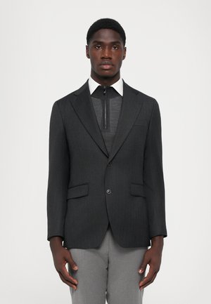 Blazer gris charbon avec des rayures verticales, deux poches avant, associé à un pull zippé gris et un col blanc, porté avec un pantalon gris clair.