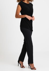 Top negro sin mangas con un diseño frontal retorcido, combinado con pantalones negros a medida y sandalias de tacón alto negras sobre un fondo blanco.