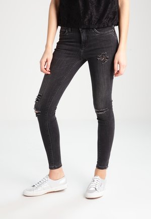 Persoon draagt zwarte gescheurde skinny jeans, zwart topje en zilveren lage sneakers en staat op een witte vloer.